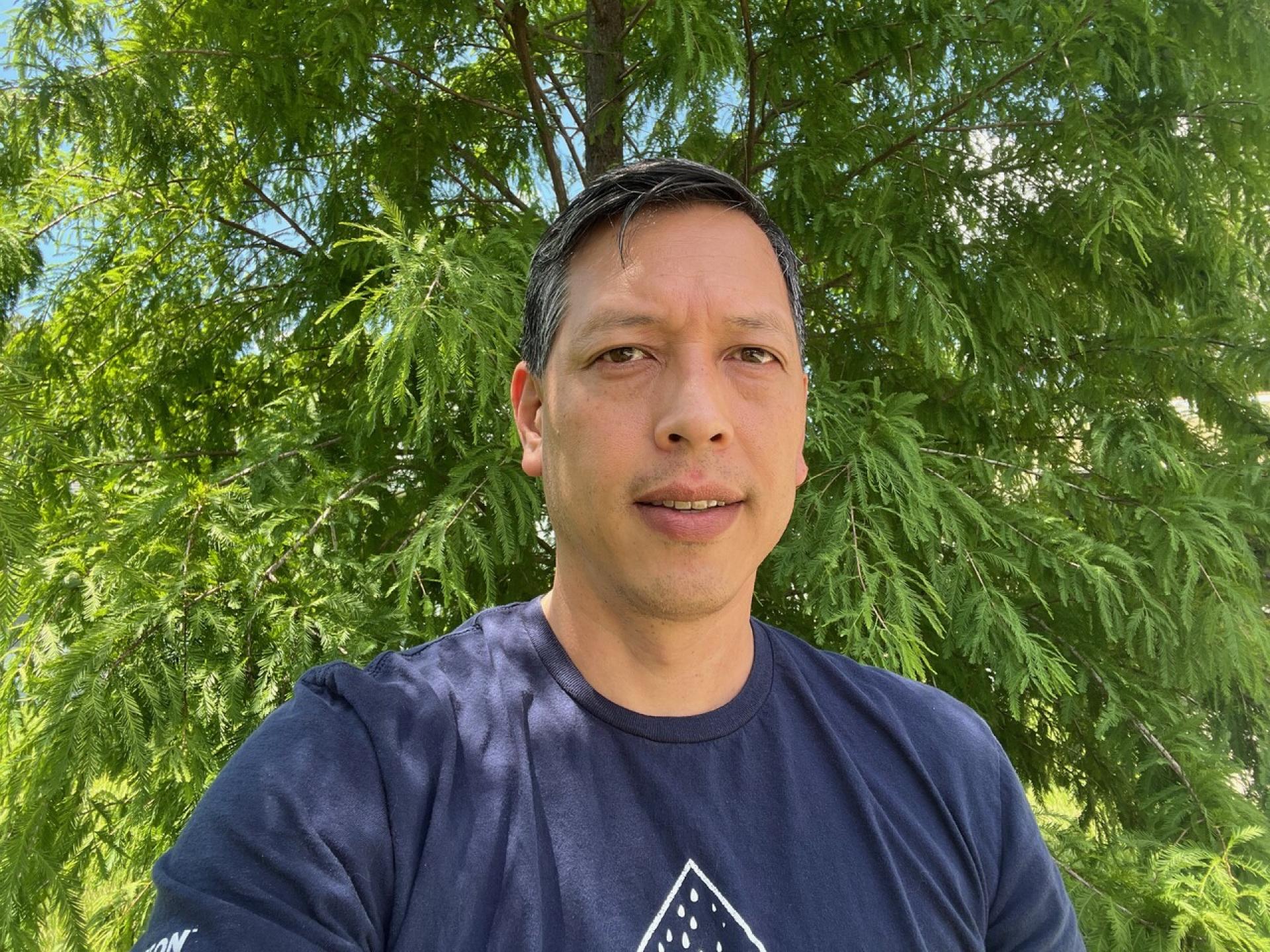 Close-up outdoor portrait of Darren Oh wearing a dark blue shirt in front of green trees.