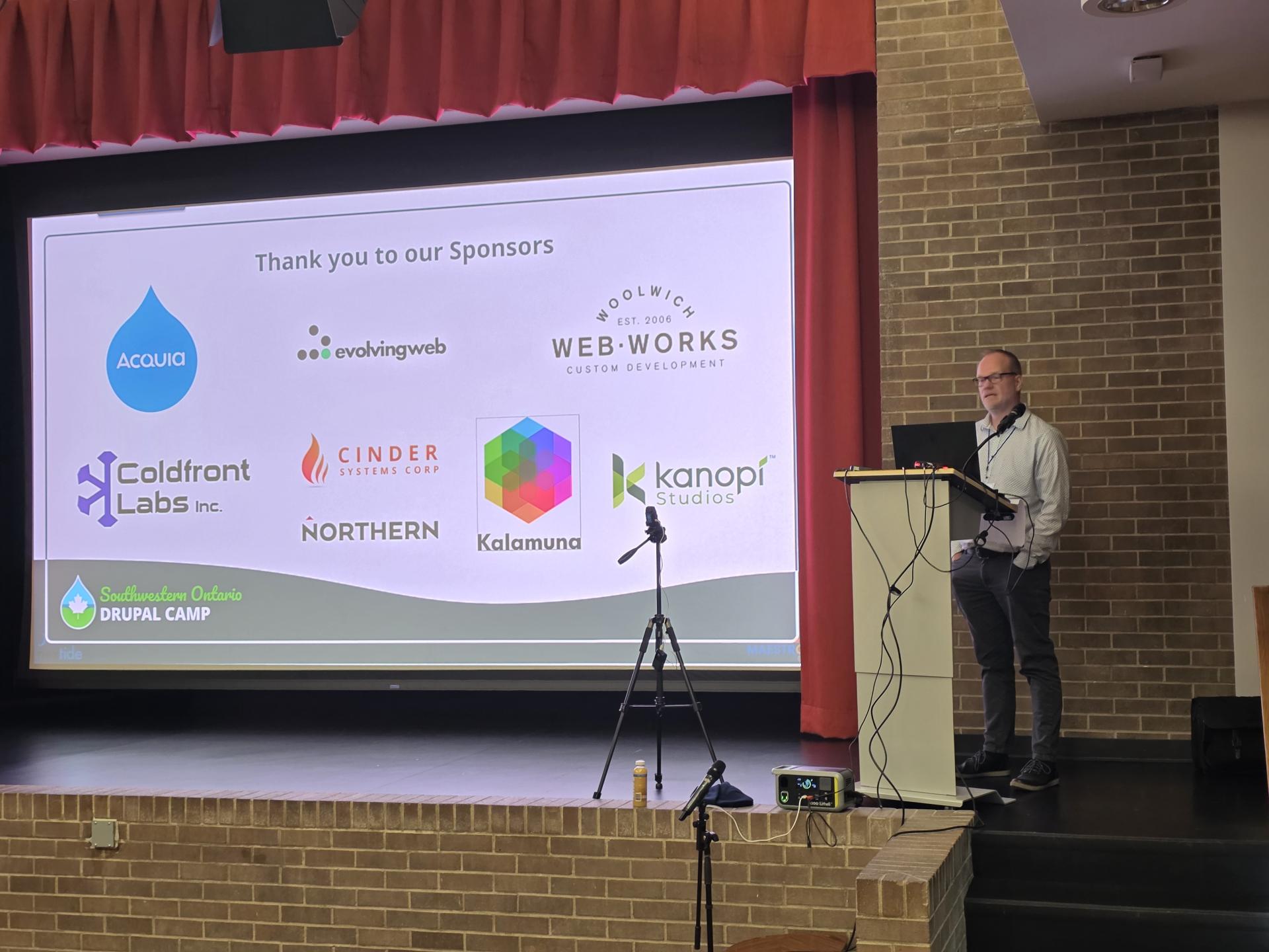 Randy stands at a podium on a brick stage beside a projected sponsor slide for Southwestern Ontario Drupal Camp. Text reads "Thank you to our Sponsors". "Acquia". "evolvingweb". "Woolwich Web Works Custom Development". "Coldfront Labs Inc.". "Cinder Systems Corp". "Northern". "Kalamuna". "Kanopi Studios". and "Southwestern Ontario Drupal Camp".
