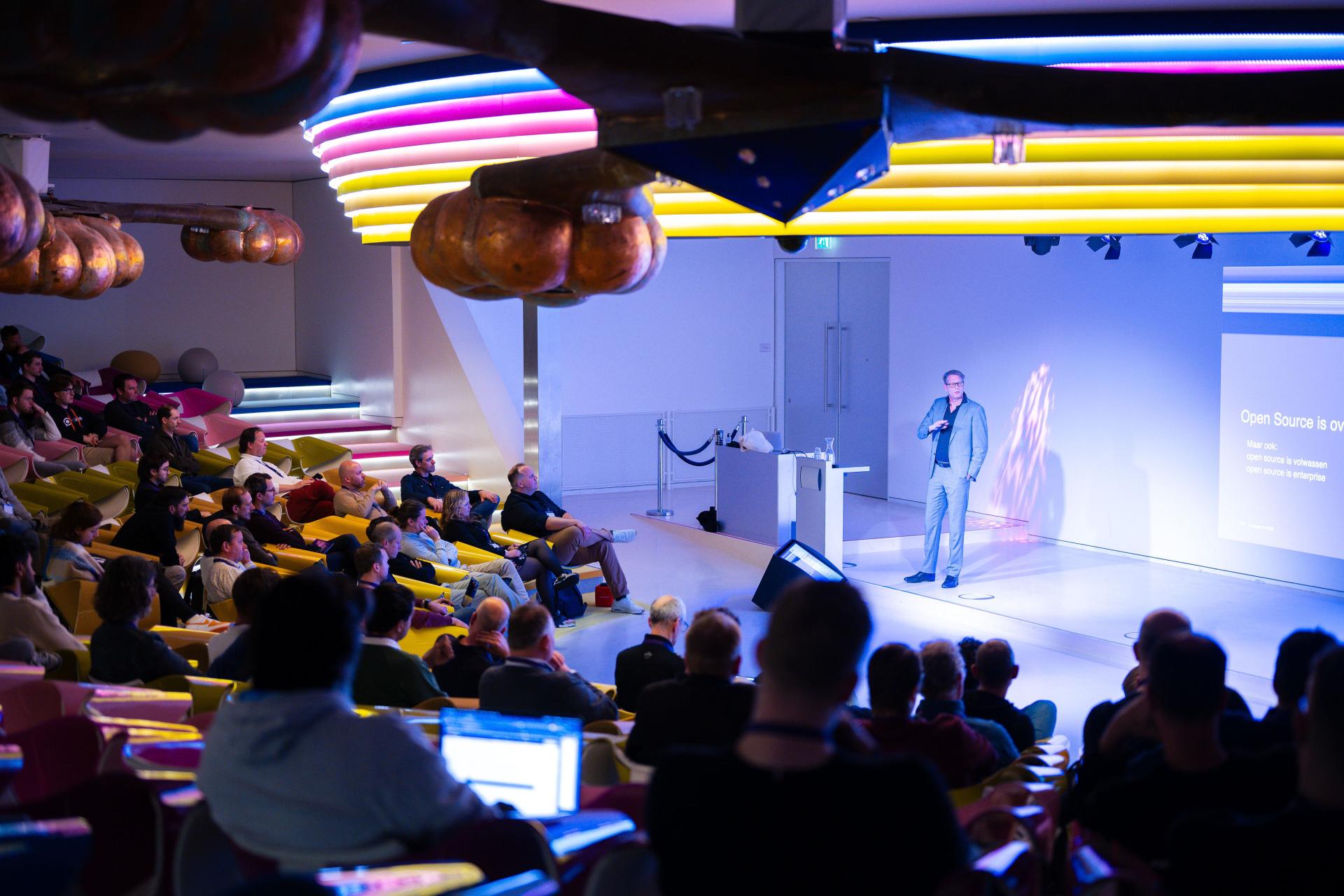 Wide photo of Bert Boerland presenting to an audience in a modern auditorium, with a projected slide visible on the right.