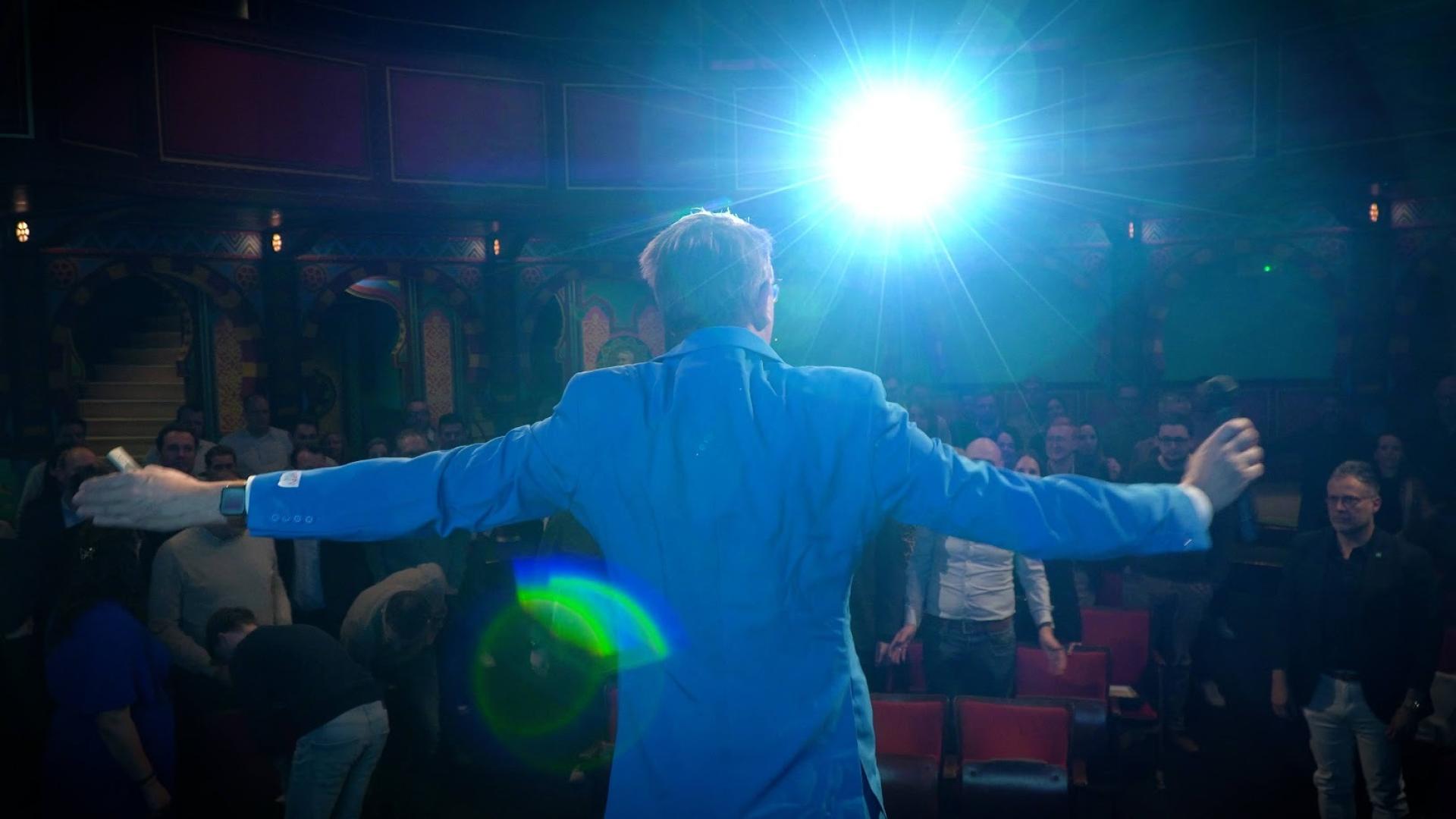 Back view of Bert Boerland standing with arms extended toward an audience at the Splash Awards venue, with a bright stage light above him.