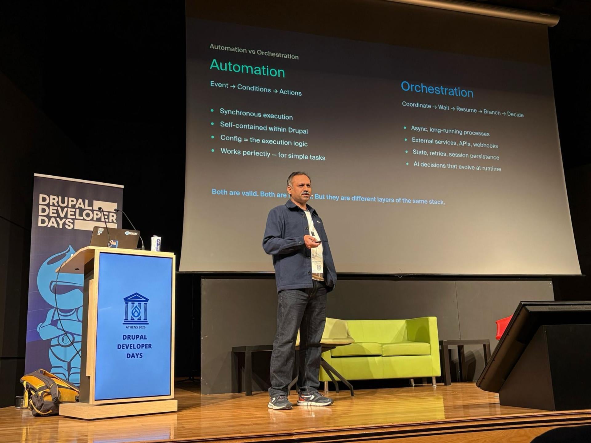 Shibin Das speaking on stage at Drupal Developer Days, with a presentation slide comparing automation and orchestration.