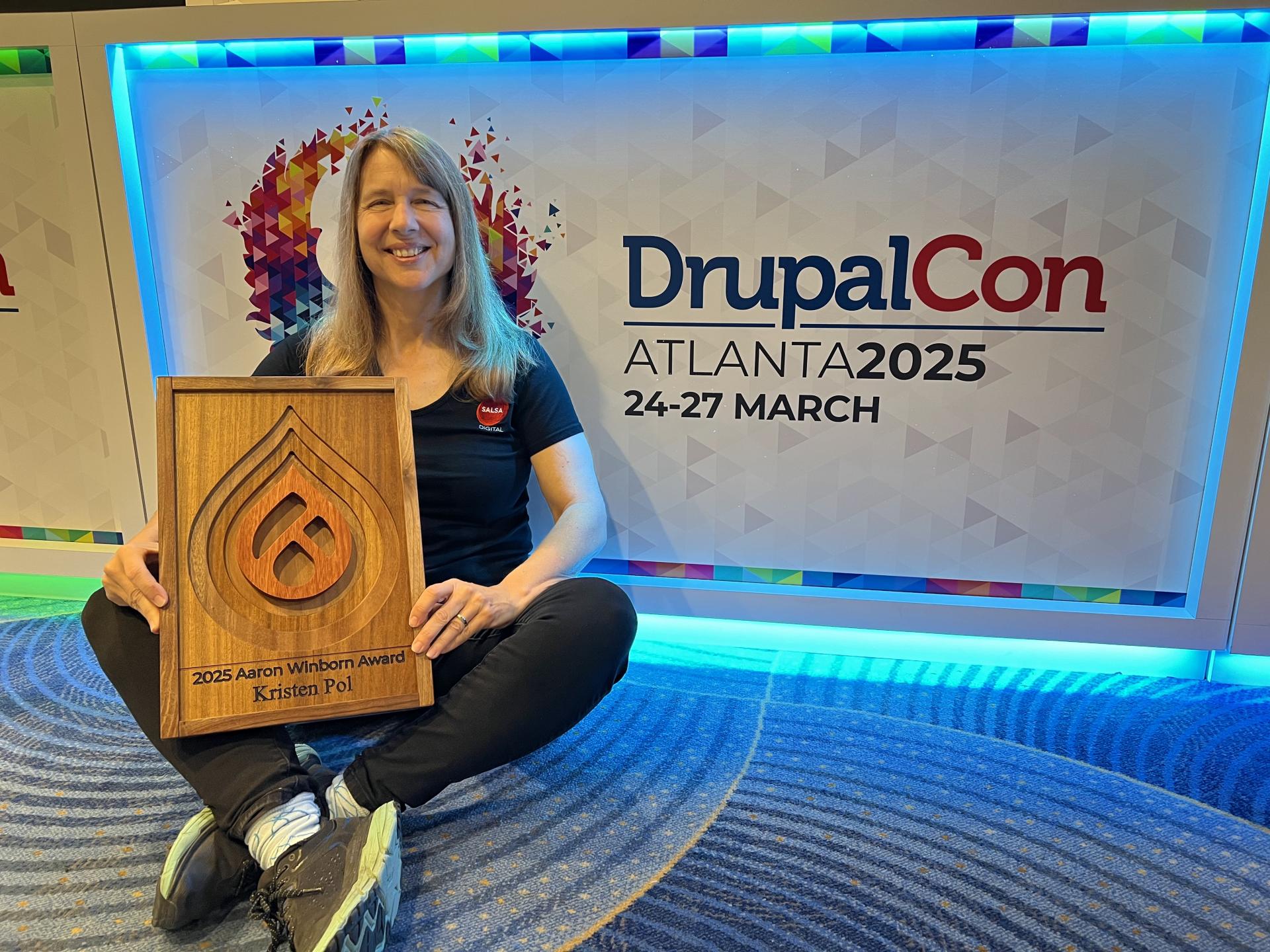 Kristen Pol seated at DrupalCon Atlanta 2025, smiling while holding the wooden 2025 Aaron Winborn Award in front of the DrupalCon Atlanta backdrop.