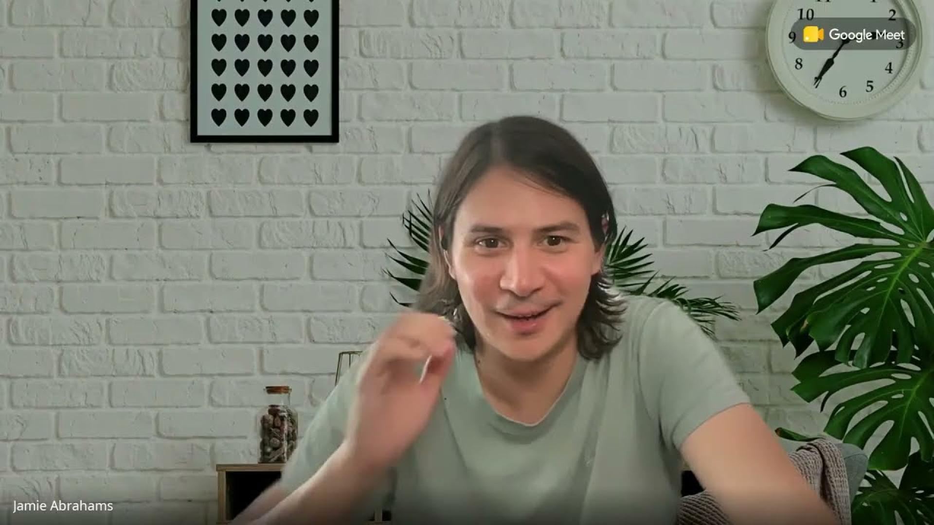 James Abrahams speaks during a Google Meet call while seated in front of a white brick wall with framed heart artwork a wall clock and large green plants. Text reads "Google Meet" and "Jamie Abrahams."