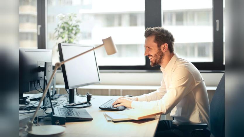 A man grinning looking at a desktop computer screen, probably chatting with hands on the keyboard