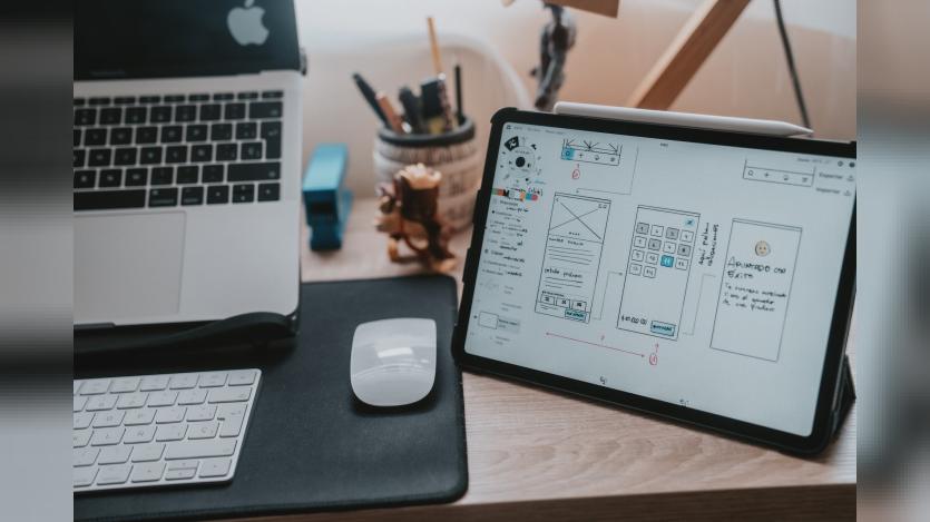 A macbook pro, mac keyboard and mouse and a tab with Ux/UI design iterations on a wooden desk