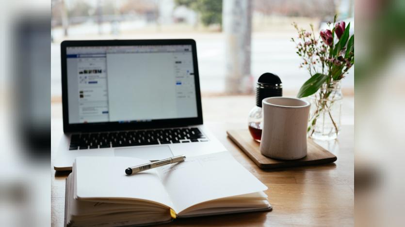 A desk with an open laptop, a notebook with pen and a cofee mug