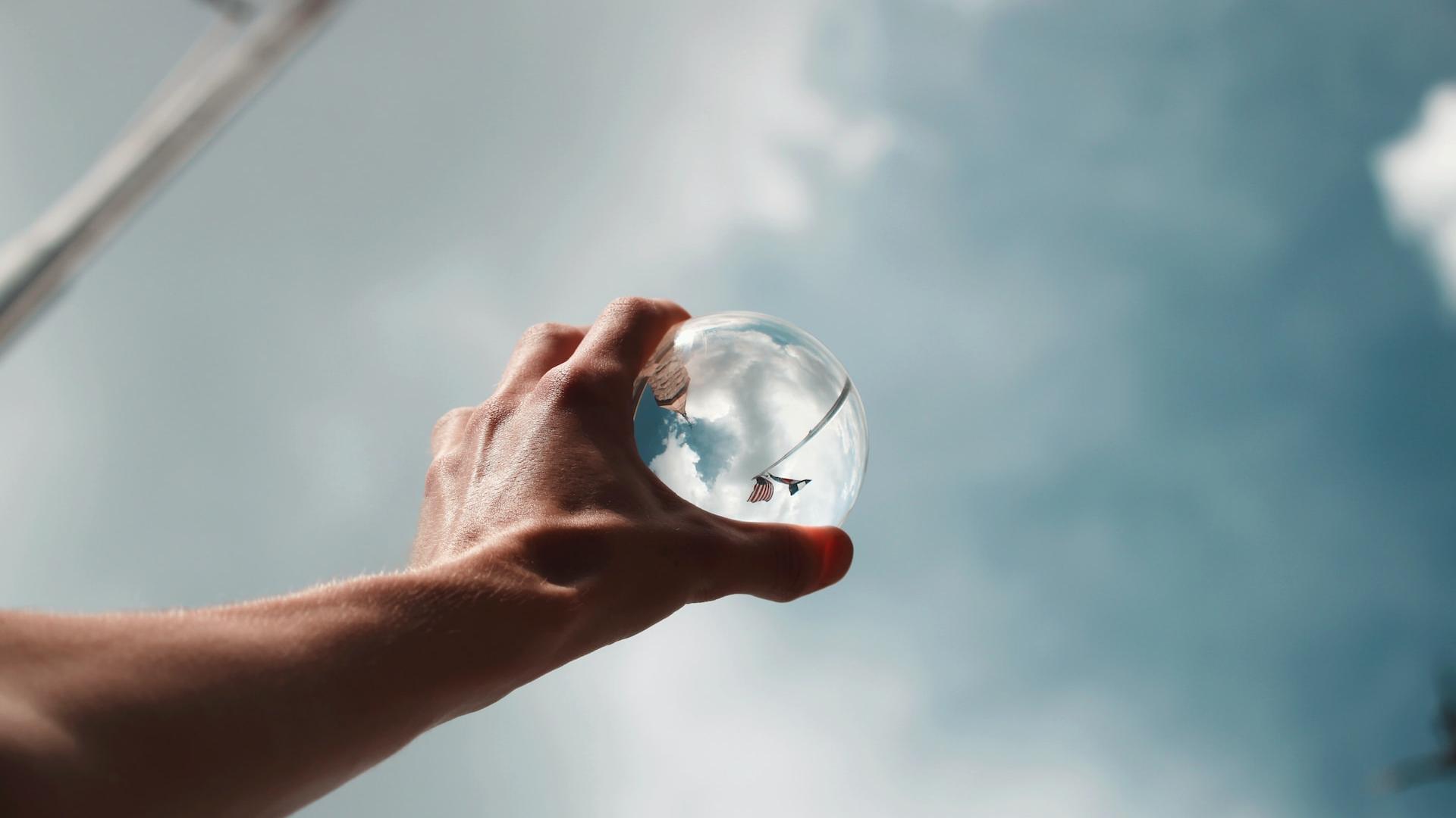 A man's hand holding a glass sphere to the skies which reflects the surroundings in a skewed manner