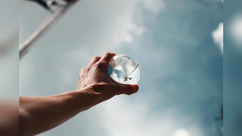 A man's hand holding a glass sphere to the skies which reflects the surroundings in a skewed manner