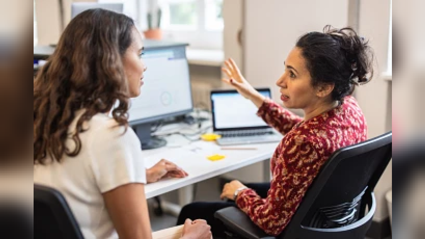 The image shows two women engaged in a discussion at a workspace, likely collaborating on a project at a desk with computer monitors.