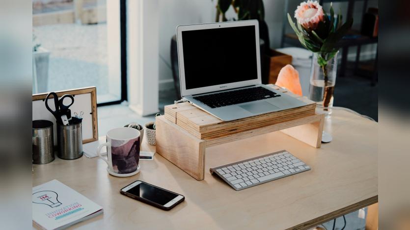 A workdesk with a laptop, wireless keyboard, mobile phone, a book, teacup, pencil holder, flowervase and other things near a glass window