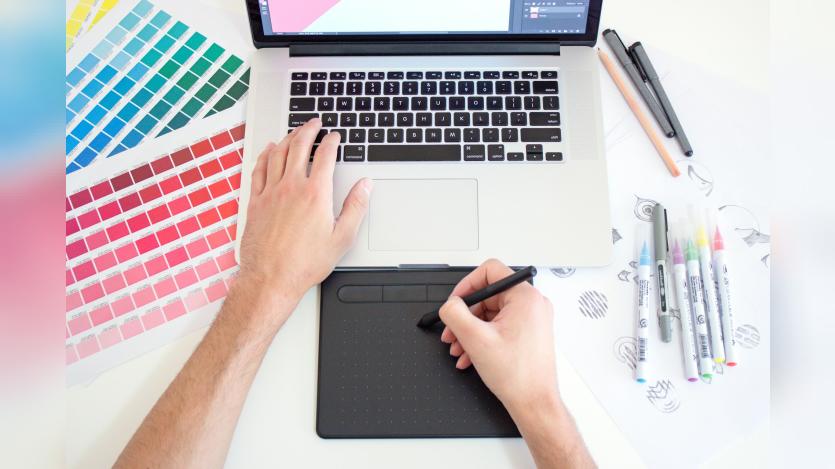 Graphic designer working on a Macbook laptop using a trackpad, color charts and markers
