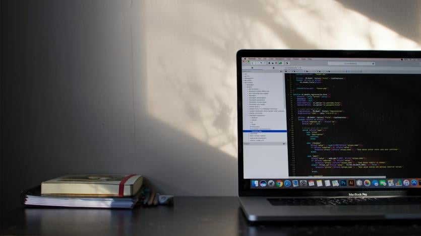 Light falling on a desk from an unseen window, a laptop with code and two books are placed on the table.