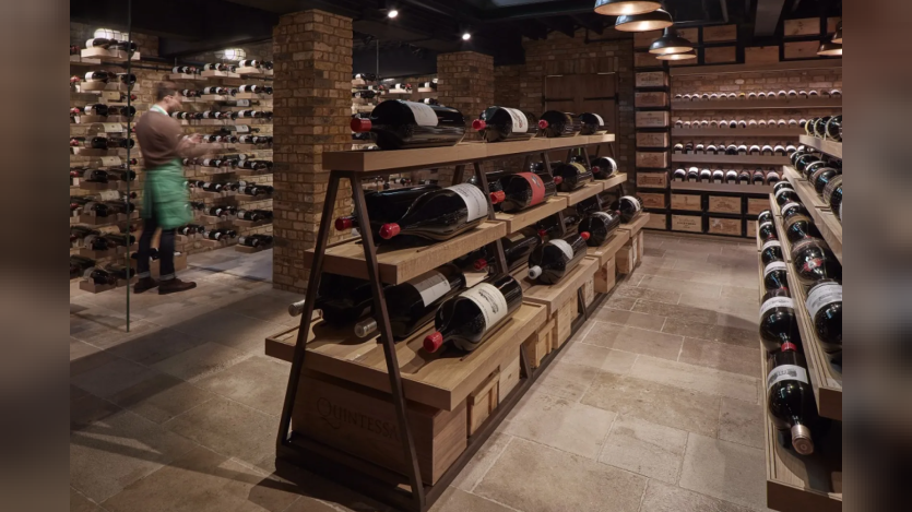 A well-lit, rustic wine cellar with exposed brick columns and a stone tile floor. In the foreground, a multi-tiered wooden A-frame shelf displays several large format wine bottles horizontally. To the left, a staff member in a green apron works near floor-to-ceiling glass-enclosed wine racks. Additional wine shelving and wooden crates line the back wall under warm overhead pendant lighting.