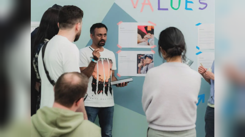 Earlier photo of Shibin Das in a Factorial team discussion, holding a tablet while several participants face posted materials on a wall marked “Values.”