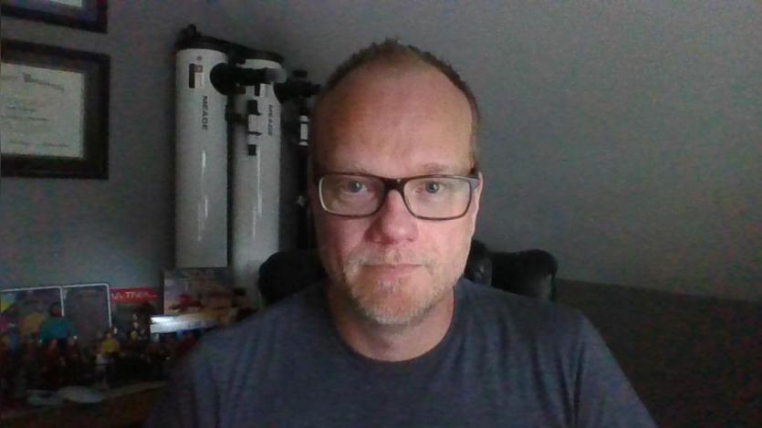 Randy wearing glasses and a dark T shirt sits facing the camera in a dim home office. Framed certificates hang on the wall behind him beside two tall apparatus and a shelf with small collectibles.