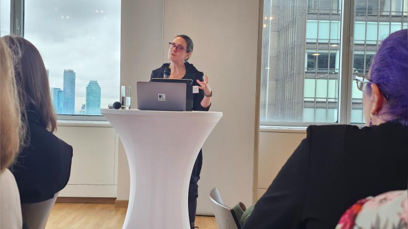 Tiffany Farris speaks into a microphone from a podium with a laptop and tablet while addressing an audience in a bright conference room. Large windows behind her overlook nearby city buildings and frame the professional presentation setting.