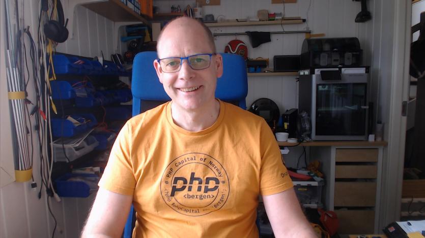 Helge Notø smiles at the camera while seated in a blue office chair inside a workshop filled with cables tools storage bins shelves and equipment. He wears blue framed glasses and an orange shirt with text reading "PHP Capital of Norway" and "php bergen."