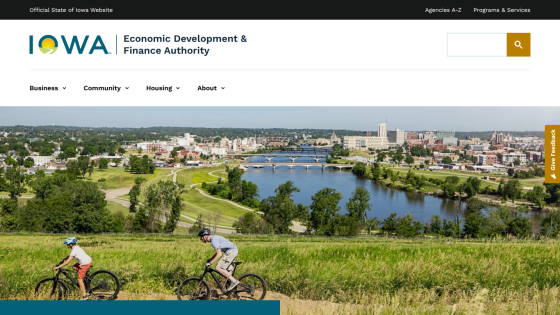 Iowa Economic Development & Finance Authority website with navigation and a hero image of two cyclists riding on a grassy hill overlooking a river and cityscape