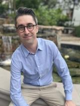 Nathan, wearing round glasses and a light blue button-down shirt with khakis, sitting on the edge of a concrete outdoor fountain with small waterfalls in the background.