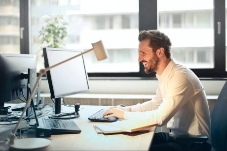 A man grinning looking at a desktop computer screen, probably chatting with hands on the keyboard