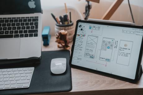 A macbook pro, mac keyboard and mouse and a tab with Ux/UI design iterations on a wooden desk