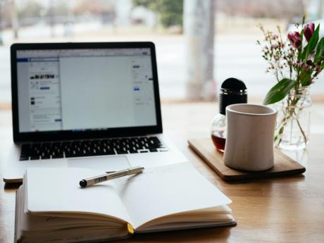 A desk with an open laptop, a notebook with pen and a cofee mug