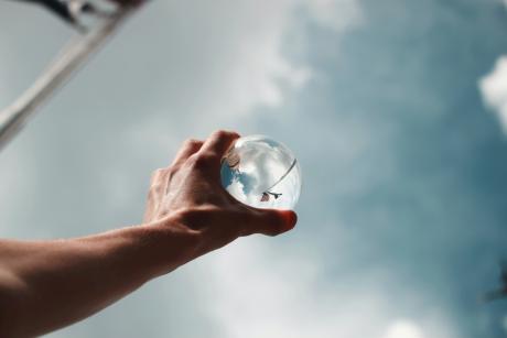 A man's hand holding a glass sphere to the skies which reflects the surroundings in a skewed manner