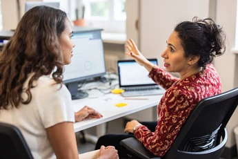 The image shows two women engaged in a discussion at a workspace, likely collaborating on a project at a desk with computer monitors.