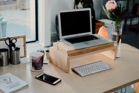A workdesk with a laptop, wireless keyboard, mobile phone, a book, teacup, pencil holder, flowervase and other things near a glass window