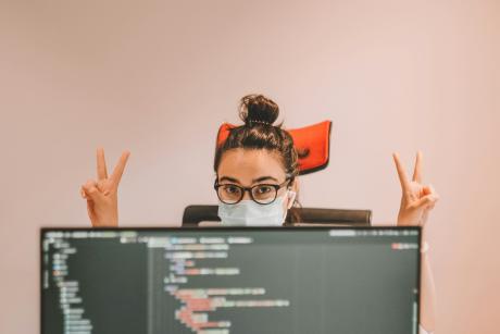 A girl wearing surgical mask behind a computer screen with victory sign in her hands