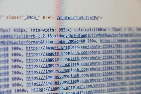 Close-up photograph of HTML and image URL code displayed on a computer screen, showing multiple links to images hosted on Unsplash. The code includes attributes such as width settings, formatting parameters, and image source URLs, with parts of the text slightly blurred.