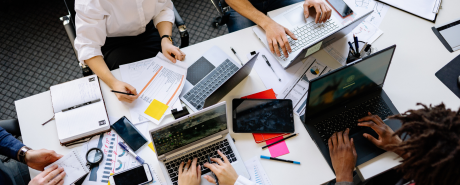 An overhead shot of a group of colleagues collaborating around a white conference table cluttered with laptops, smartphones, notebooks, pens, and printed charts and documents.