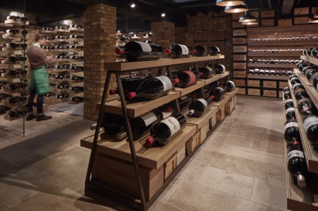 A well-lit, rustic wine cellar with exposed brick columns and a stone tile floor. In the foreground, a multi-tiered wooden A-frame shelf displays several large format wine bottles horizontally. To the left, a staff member in a green apron works near floor-to-ceiling glass-enclosed wine racks. Additional wine shelving and wooden crates line the back wall under warm overhead pendant lighting.