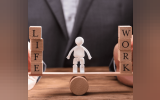 Demonstrating Work Life Balance using a tiny wooden seesaw with wooden blocks on either side and a small white human effigy on the center