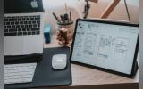 A macbook pro, mac keyboard and mouse and a tab with Ux/UI design iterations on a wooden desk