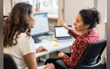 The image shows two women engaged in a discussion at a workspace, likely collaborating on a project at a desk with computer monitors.