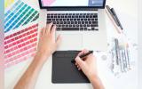 Graphic designer working on a Macbook laptop using a trackpad, color charts and markers