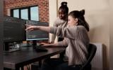 Caucasian network developer pointing out high usage of processing power in mainframe grid. African american programmer helping coworker to solve overloaded system storage space.