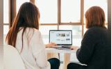 Two women seen from behind, seated on either side of a laptop and facing the same screen.